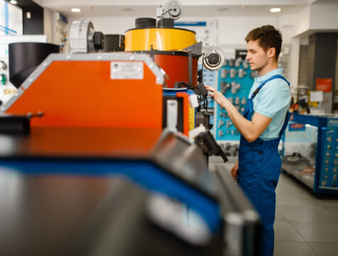 Plumber in uniform choosing boiler with furnace at showcase in plumbering store. Man with notebook buying sanitary engineering in shop, water heater choice